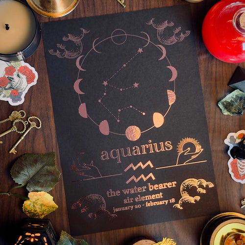 A flat-lay of a black astrology print featuring the Aquarius zodiac sign in rose gold foil, surrounded by mystical props like a candle, brass candlestick, keys, crystals, and dried leaves on a dark wooden table.