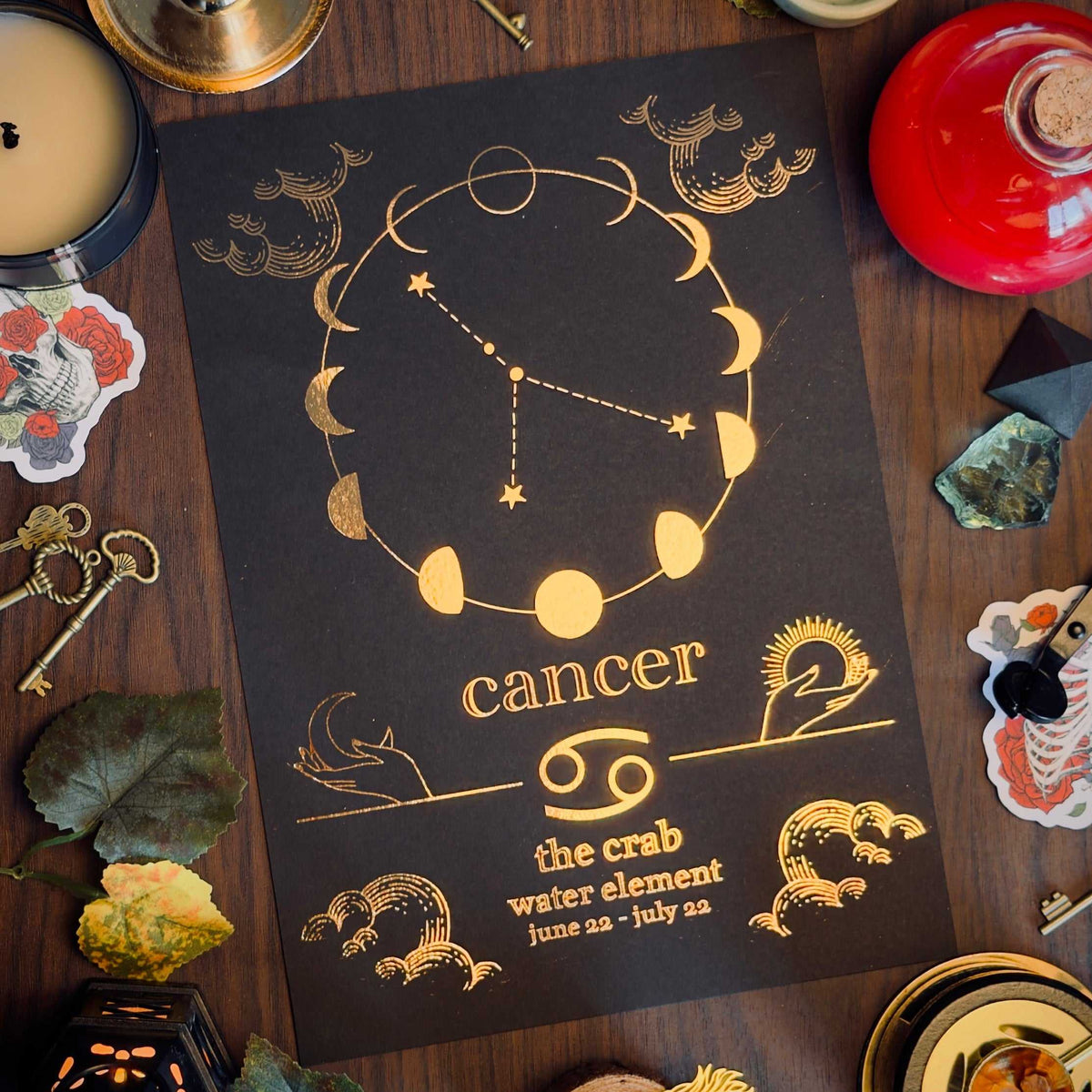 A black Cancer zodiac print with gold celestial graphics and text, styled in a flat lay on a dark wooden table with candles, keys, leaves, crystals, and decorative accents.