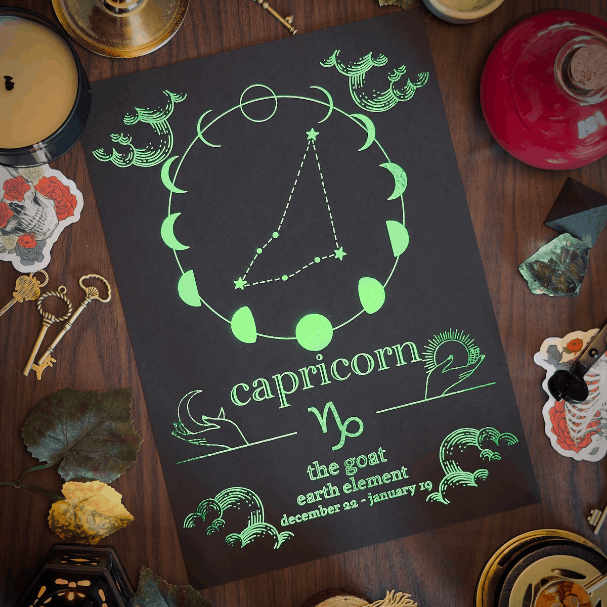 A flat-lay of a black card featuring a green astrological chart for Capricorn, surrounded by mystical props like a candle, keys, crystals, and a red glass bottle on a dark wooden table.
