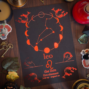 A square black card or print featuring orange astrological illustrations and text for Leo, including moon phases, a lion silhouette, and the phrase 'the lion fire element' on a dark wood surface surrounded by decorative props.