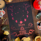 A Libra-themed invitation card or print laid flat on a dark wooden table, surrounded by candles, keys, crystals, leaves, and small decorative objects. The card is black with pink astrological line art and the word 'libra' centered, with additional text below.