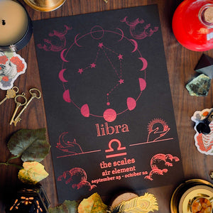 A Libra-themed invitation card or print laid flat on a dark wooden table, surrounded by candles, keys, crystals, leaves, and small decorative objects. The card is black with pink astrological line art and the word 'libra' centered, with additional text below.