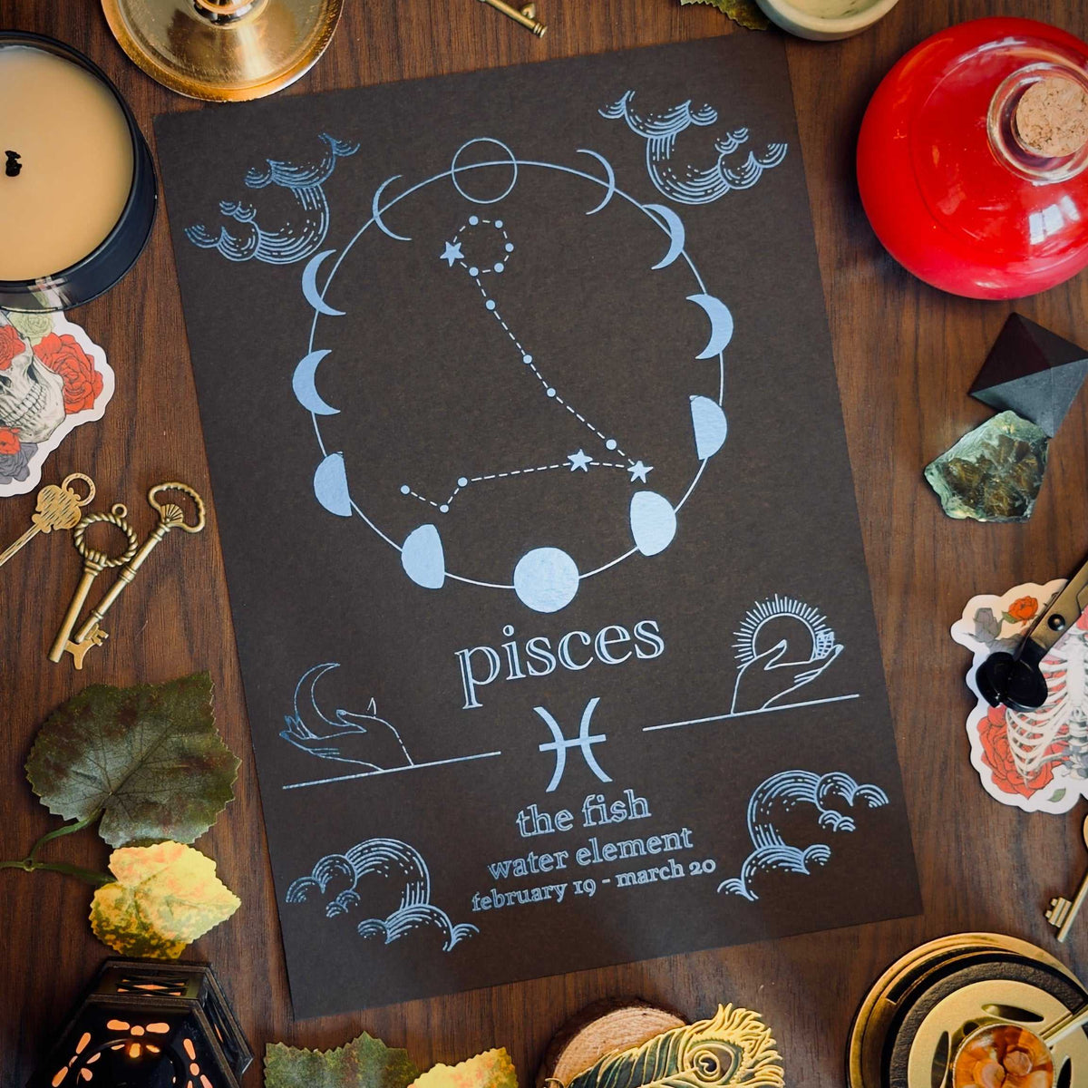 A tarot card or astrology print featuring Pisces with moon phases, fish motifs, and the text 'the fish water element' is displayed on a dark wooden table surrounded by candles, keys, crystals, and autumn leaves.