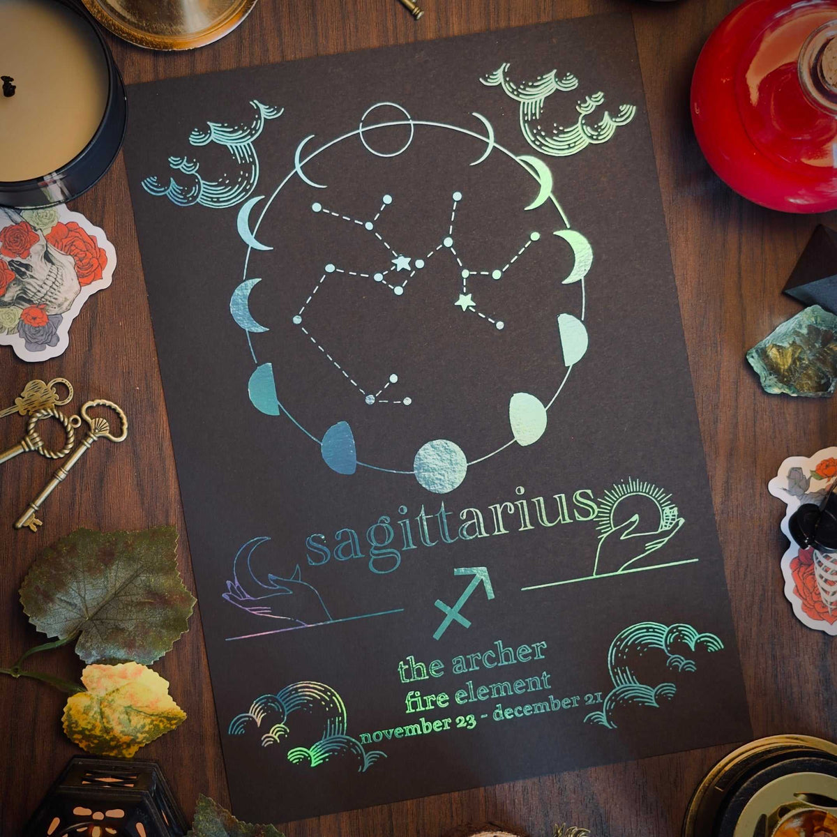 A dark tarot-style card featuring the Sagittarius constellation and moon phases, styled on a wooden surface with candles, crystals, keys, and dried leaves.