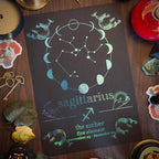 A dark tarot-style card featuring the Sagittarius constellation and moon phases, styled on a wooden surface with candles, crystals, keys, and dried leaves.