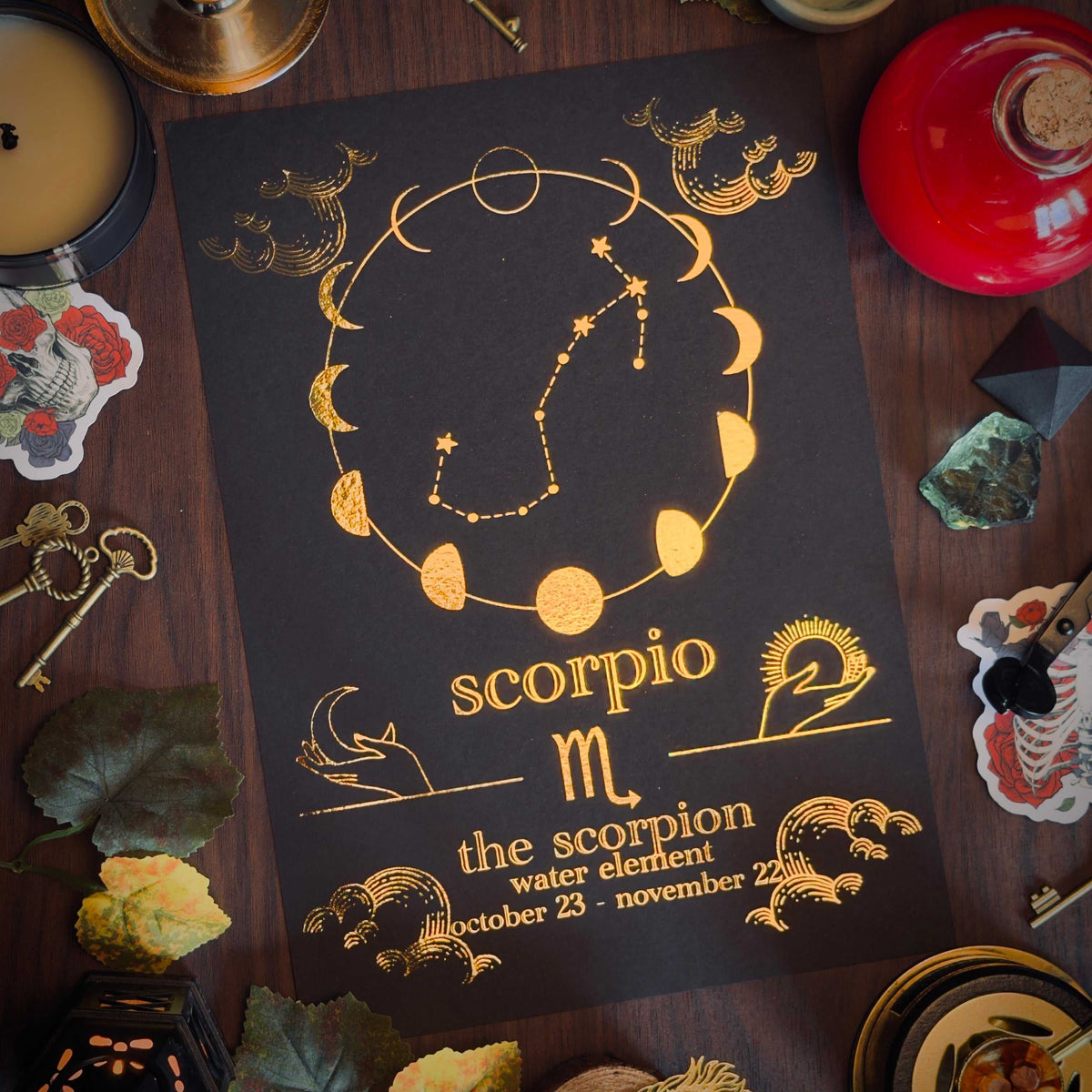 A black astrology card featuring gold foil artwork of the Scorpio zodiac sign, surrounded by mystical props like crystals, a candle, and a red glass bottle on a dark wooden surface.