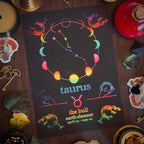 A black zodiac art print for Taurus, featuring a colorful retro constellation design and text, styled flat-lay on a dark wooden table surrounded by crystals, candles, and decorative items.