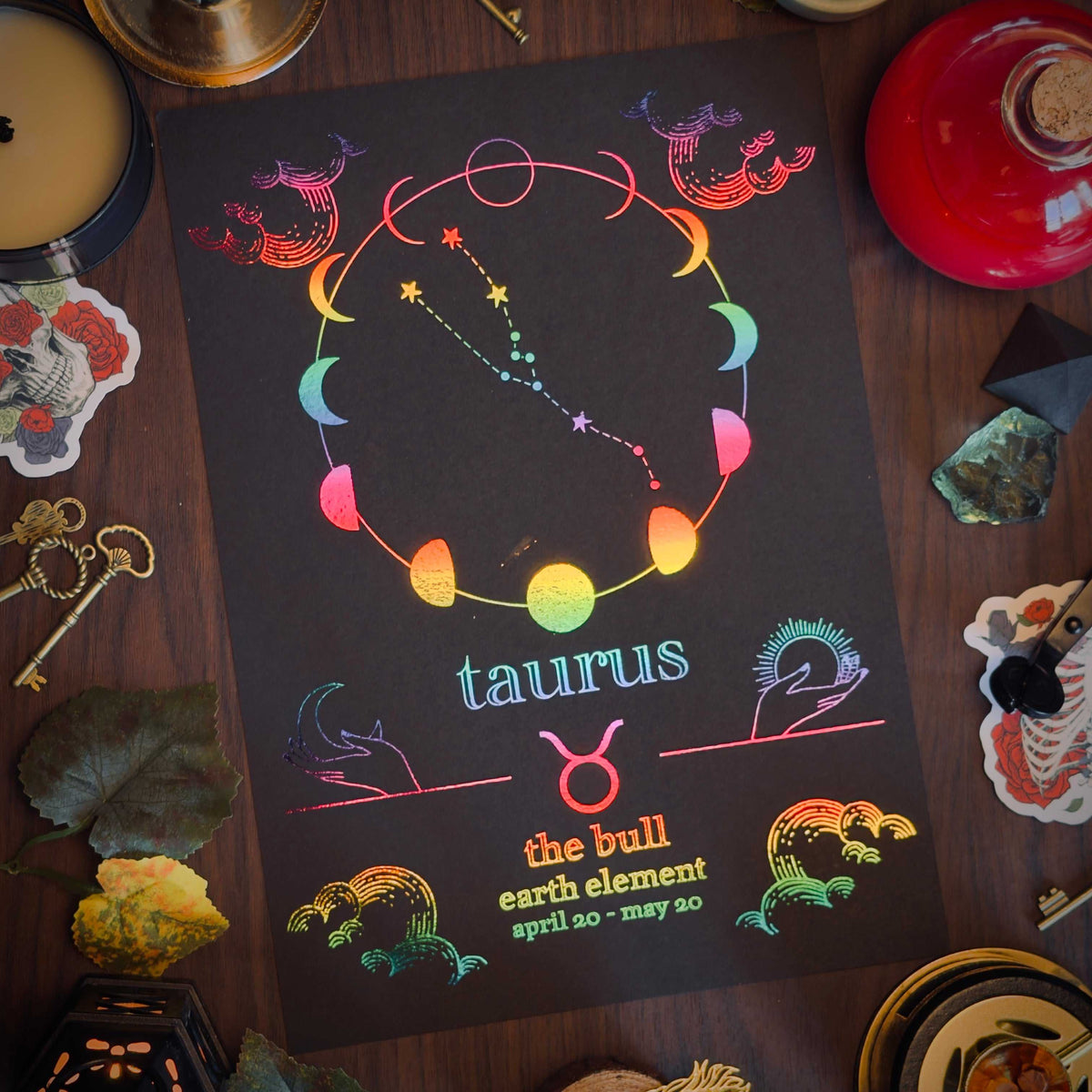 A black zodiac art print for Taurus, featuring a colorful retro constellation design and text, styled flat-lay on a dark wooden table surrounded by crystals, candles, and decorative items.