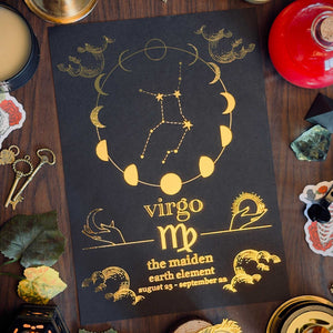 A black astrology print featuring the Virgo constellation and moon phases in gold, styled in a flat lay on a dark wooden table with candles, keys, crystals, and leaves.