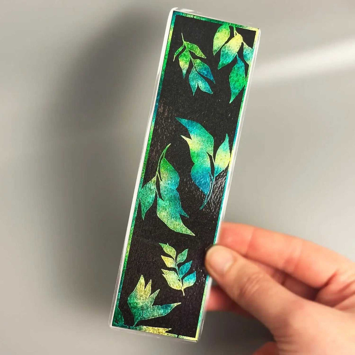Hand holding a rectangular bookmark with vibrant green and blue leaf patterns on black background