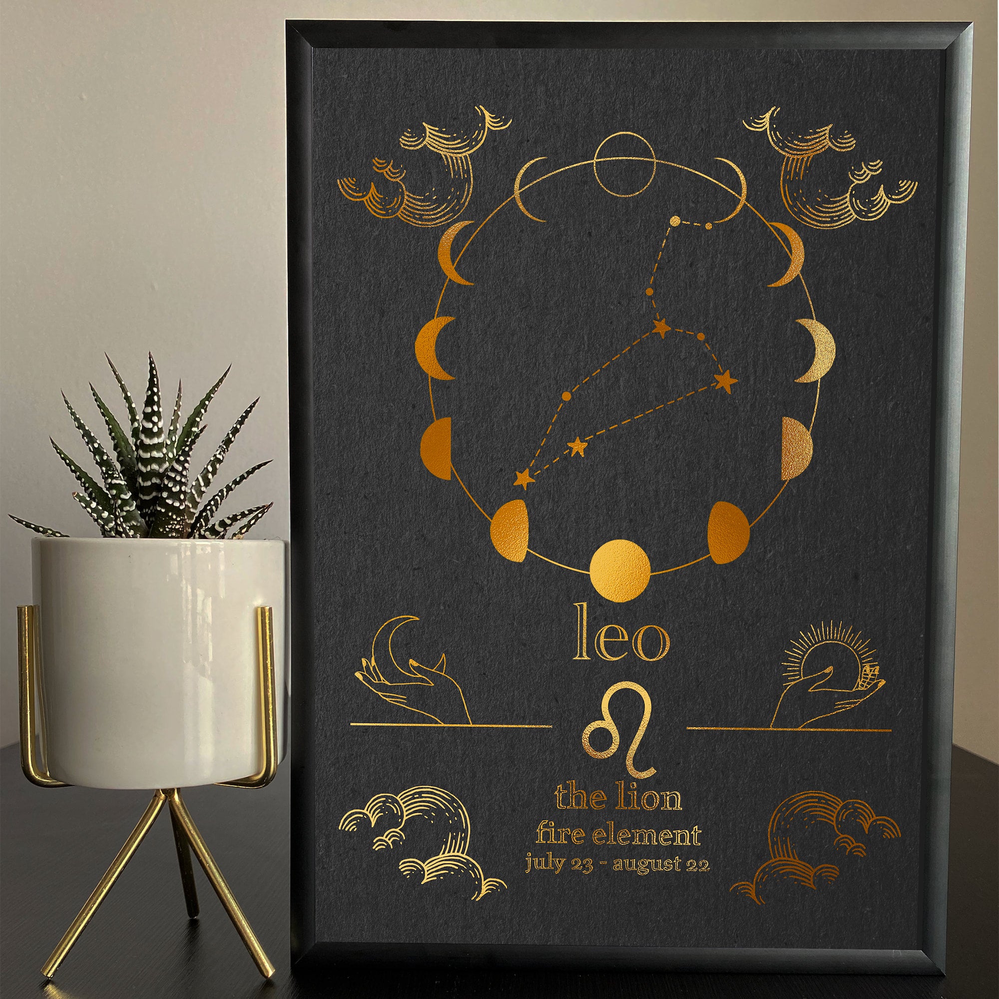 A framed artwork featuring the Leo zodiac sign with a foil finish, displayed on a table with a plant and decorative items.