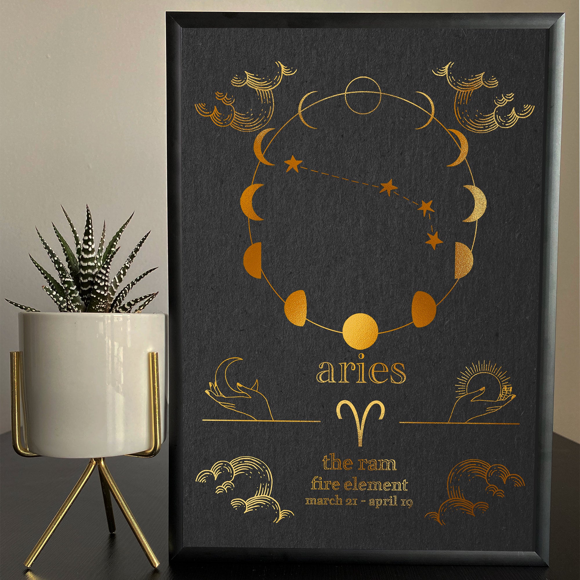 A framed Aries zodiac-themed artwork with a foil finish, featuring the constellation and attributes of the Aries sign, displayed next to a potted plant.