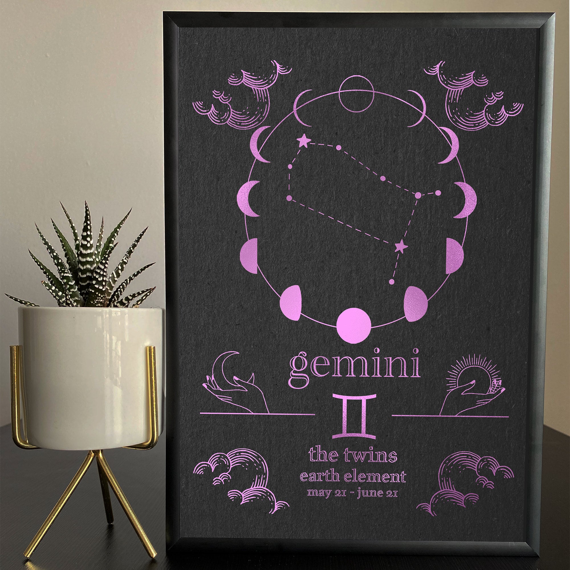 A framed foil print featuring the Gemini zodiac sign with a pink and black color scheme, displayed on a table with a plant and decorative items.